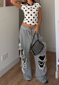 Woman wearing a white cropped polka dot shirt and ripped grey wide-leg trousers, holding a black studded handbag indoors.