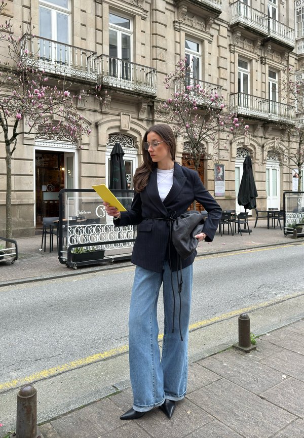Blazer nero a righe con design avvolgente, t-shirt bianca, jeans larghi azzurri chiari, borsa nera, occhiali trasparenti e bloc-notes giallo.