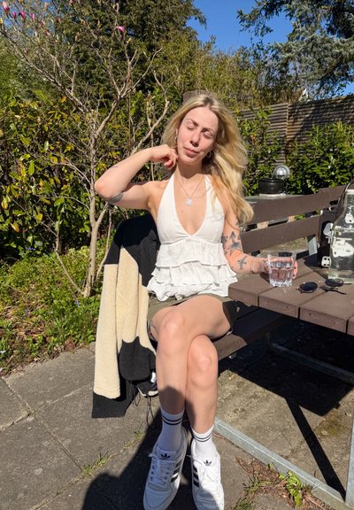 Mujer con camiseta blanca sin mangas y shorts sentada en un banco al aire libre bajo la luz del sol, sosteniendo un vaso de agua con una chaqueta apoyada cerca.