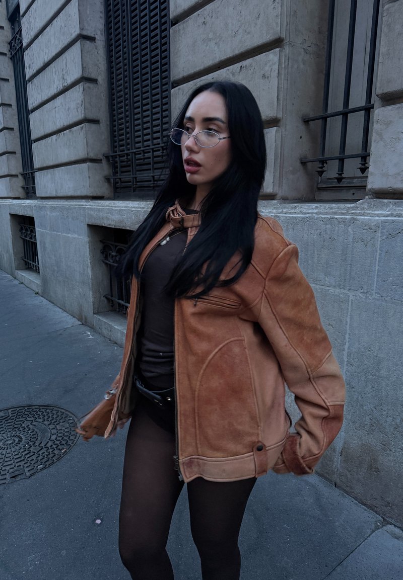 Vrouw met lang zwart haar en een bril, met een bruine jas en een donker tenue, staande op een stoep in de stad nabij een stenen gebouw.