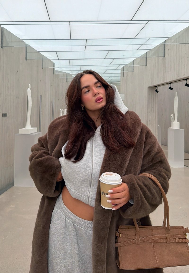 Brown faux fur coat over a grey zip-up hoodie and matching joggers. Holding a coffee cup and a brown suede handbag. Minimalist background.