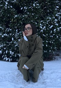 Veste d'extérieur verte surdimensionnée avec de gros boutons, tissu texturé et col montant. Portée par-dessus une chemise blanche à manches longues, assise dans la neige.