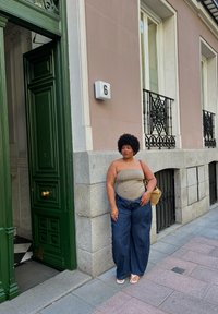 Žmogus stovi prie žalių durų, dėvi šviesiai rudą liemenę ir plačius mėlynus džinsus, laiko smėlio spalvos krepšį, avėdamas baltus sandalus ant šaligatvio.