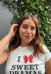 Hvid t-shirt med sort tekst, der læser "I SWEET DRAMAS" og en rød sløjfe grafisk. Baggrunden viser grønne efeu blade. En person rører ved håret.