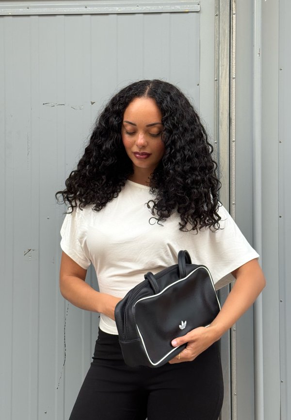 Femme aux cheveux bouclés portant une chemise blanche et un pantalon noir, tenant un sac noir Adidas devant un fond métallique gris.