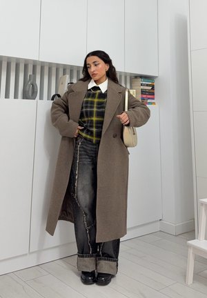 Abrigo largo de lana marrón con solapas extragrandes, superpuesto a un suéter a cuadros negro y amarillo. Combinado con jeans oscuros desgastados y zapatos negros.