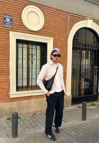 Persona vestita con una camicia bianca a righe, pantaloni neri e sneakers si trova davanti a un muro di mattoni, con una borsa a tracolla nera e occhiali da sole.