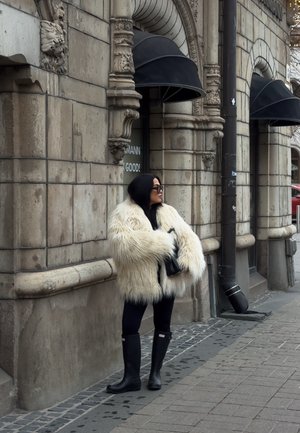 Mujer con abrigo de piel blanco sobredimensionado y botas negras de pie en la acera junto a un edificio de piedra ornamentado con toldos negros.
