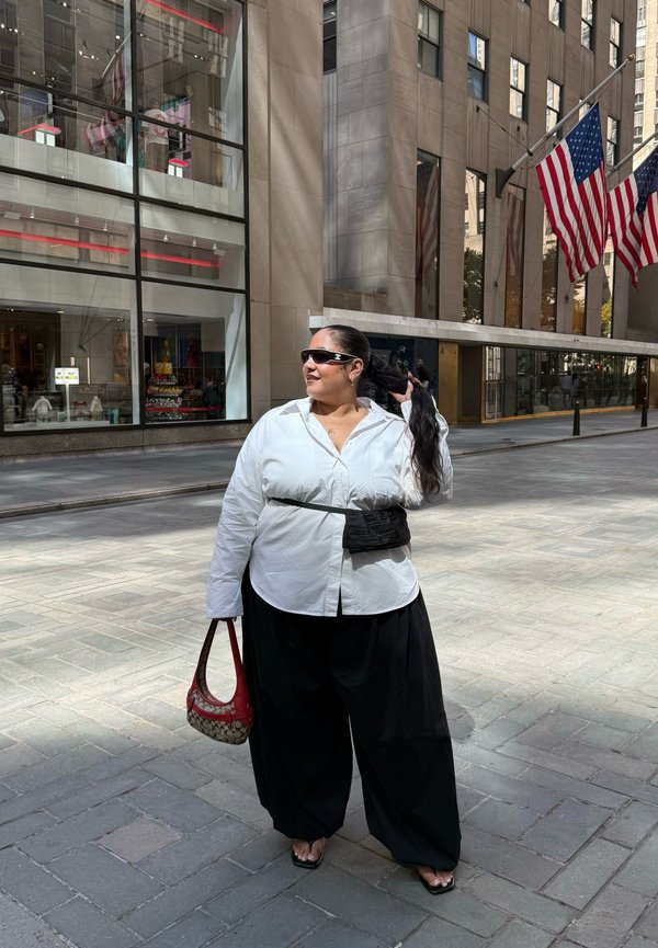 Frau in weißem Hemd und schwarzen Hosen hält eine rote Handtasche und richtet ihre Haare auf einer Stadtstraße mit amerikanischen Flaggen und einem Glasgebäude im Hintergrund.