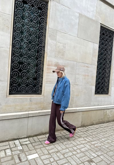 Chaqueta bomber de mezclilla, suéter corto marrón, pantalones de chándal burdeos con rayas blancas y chanclas rosas. Rejas de ventana en espiral negras de fondo.