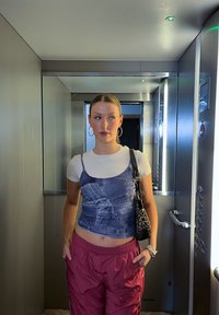 Νεαρή γυναίκα με σκουλαρίκια κρίκους, φορώντας στρωτές λευκές και τζιν μπλούζες, κόκκινο παντελόνι και τσάντα με μοτίβο λεοπάρ, στέκεται σε μεταλλικό ανελκυστήρα.