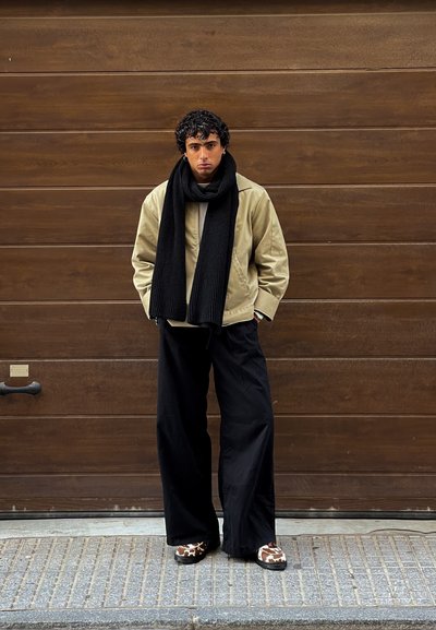 Chaqueta beige, bufanda negra de canalé, parte superior clara, pantalones negros anchos, zapatos marrones y blancos con un estampado de vaca. De pie contra un fondo de madera.