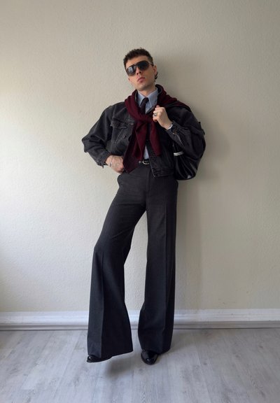 Persona posando en interiores vistiendo pantalones de pierna ancha con rayas finas, chaqueta de mezclilla negra, suéter burdeos sobre los hombros, corbata, gafas de sol y sosteniendo un bolso negro.