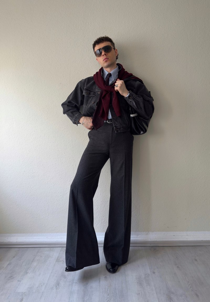 Person, die drinnen posiert, trägt weite Streifenhosen, schwarze Jeansjacke, einen burgunderfarbenen Pullover über den Schultern, Krawatte, Sonnenbrille und hält eine schwarze Handtasche.