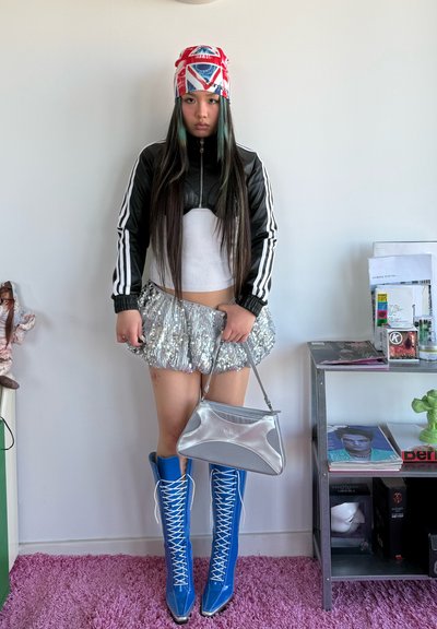 Joven con cabello largo que lleva un pañuelo rojo y blanco en la cabeza, chaqueta en blanco y negro, falda de lentejuelas plateadas, botas azul con cordones y sostiene un bolso plateado.