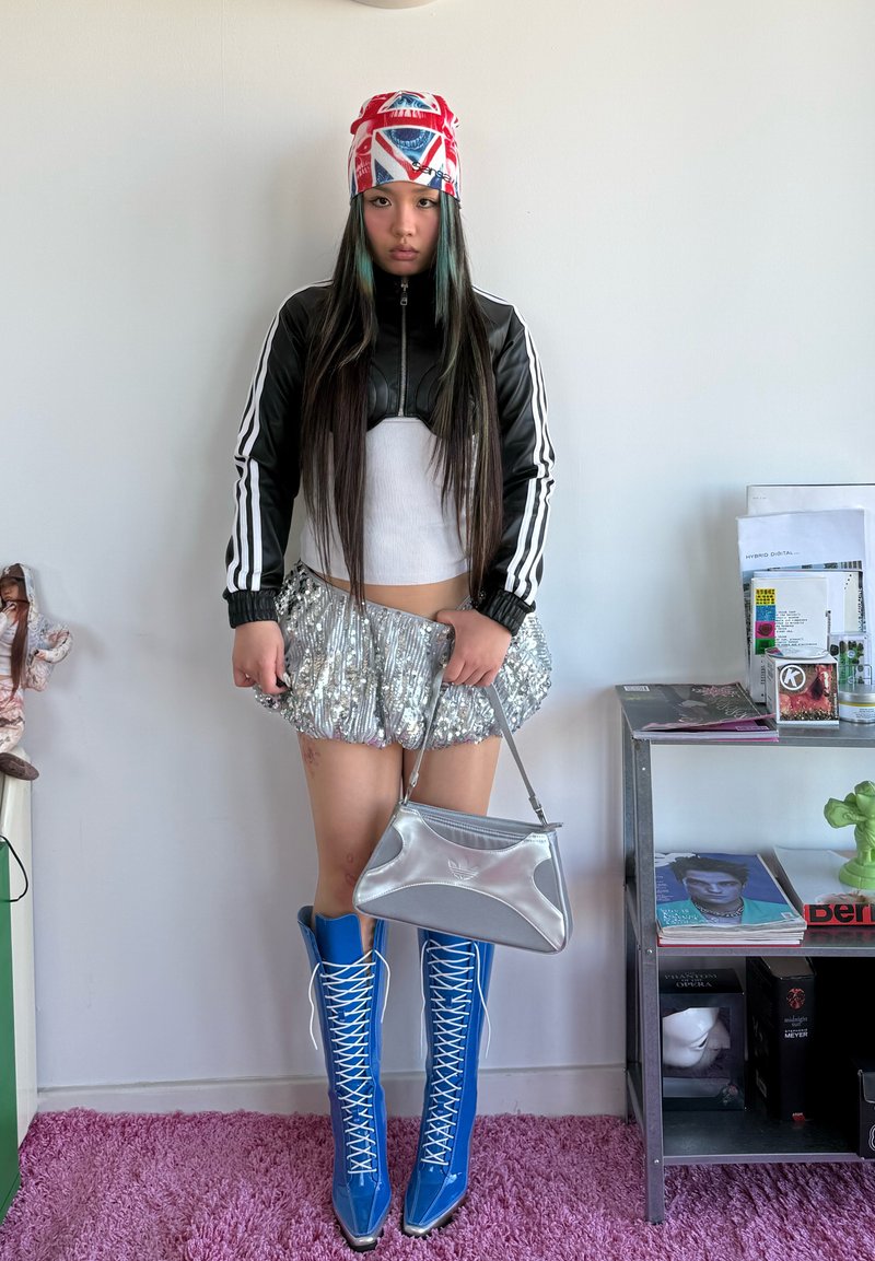 Tânără cu păr lung, purtând un batic roșu și alb, o jachetă alb-negru, o fustă argintie cu paiete, cizme albastre cu șireturi și ținând o geantă de mână argintie.