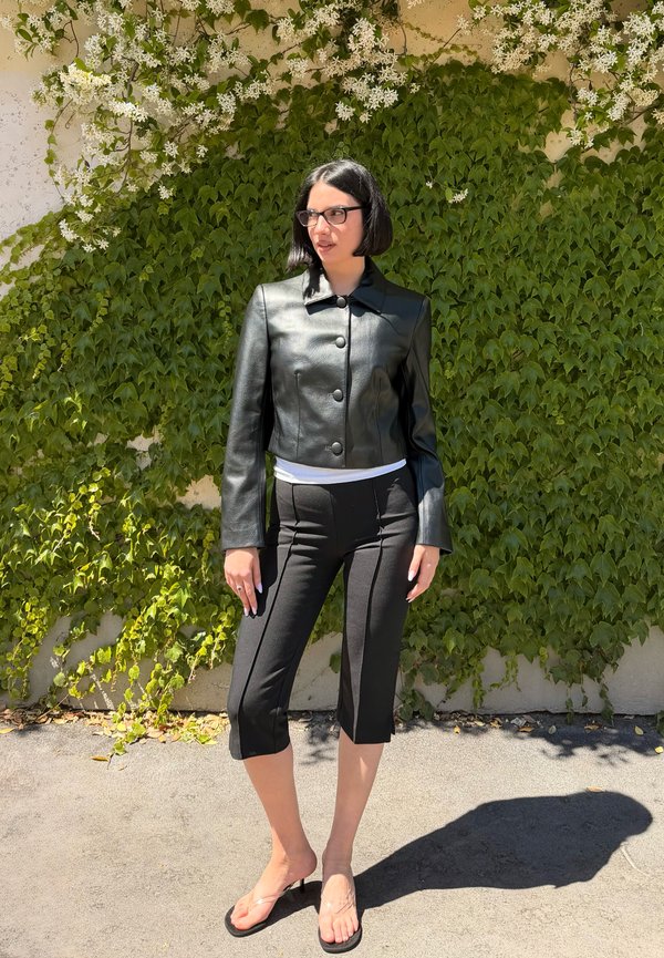 Femme en veste courte en cuir noire et pantalon capri, debout pieds nus sur un trottoir baigné de soleil devant un mur feuillu avec des fleurs blanches.