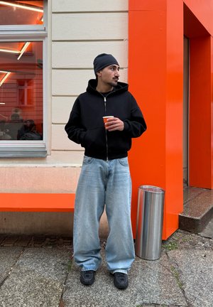 Juodas užtrauktuku užsegamas džemperis su gobtuvu, šviesiai mėlynos spalvos plačios džinsinės kelnės ir juodi batai. Laiko mažą oranžinį puodelį, stovėdamas šalia oranžinės sienos.