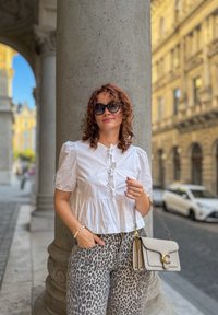 Bijela bluzica kratkih rukava s detaljima na gumbima, uparena s hlačama širokih nogavica s leopard uzorkom. Dodatno accessorirana svijetlosivom torbicom i sunčanim naočalama.