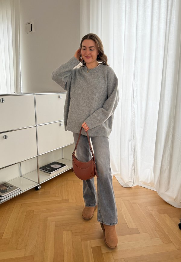 Suéter de punto gris, jeans acampanados gris claro, botas de tobillo marrones y un bolso cruzado marrón. Ambientado en un interior luminoso con suelo de madera.