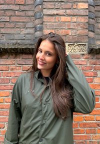 Giacca impermeabile verde oliva con chiusura a bottoni, collo alto e maniche lunghe, posta davanti a un muro di mattoni con trama. Visibili fiocchi di neve leggeri.