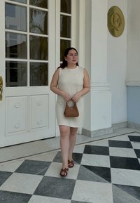 Robe sans manches beige et texturée, sac à main marron avec une poignée circulaire, sandales marron. Fond avec de grandes portes en verre et des carreaux à motifs.