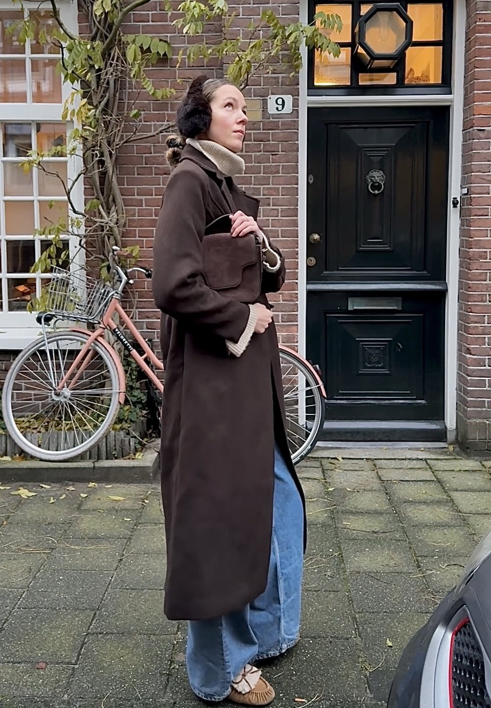 Palton lung, maro închis, cu un guler bej, asortat cu blugi albaștri, pantaloni largi, și o geantă mică maro. Cadru în aer liber, cu o bicicletă pe fundal.
