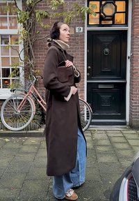 Palton lung, maro închis, cu un guler bej, asortat cu blugi albaștri, pantaloni largi, și o geantă mică maro. Cadru în aer liber, cu o bicicletă pe fundal.