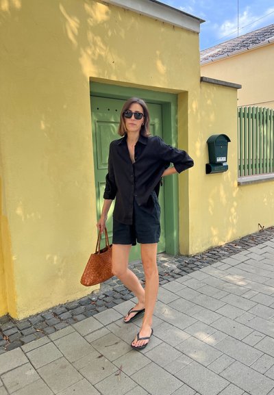 Camisa negra de botones, pantalones cortos oscuros, bolsa tejida marrón y chanclas. Puerta verde con adoquines grises. Pared amarilla de fondo.