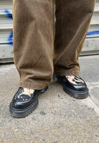 Μαύρα συνθετικά παπούτσια με υφασμάτινο σχέδιο, που διαθέτουν παχύ καουτσούκ πάτο και διακοσμητικό αντίθετο ύφασμα στο πάνω μέρος. Φοράνε καφέ παντελόνι.