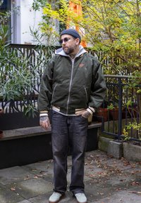 Veste bomber vert olive avec accents blancs et capuche grise, associée à un jean foncé et des baskets de couleur claire. L'arrière-plan présente de la verdure.