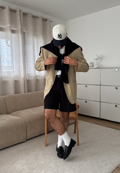 Blazer beige sobre un suéter negro caído sobre los hombros, camiseta blanca, pantalones cortos negros, calcetines blancos y mocasines negros en una habitación minimalista.