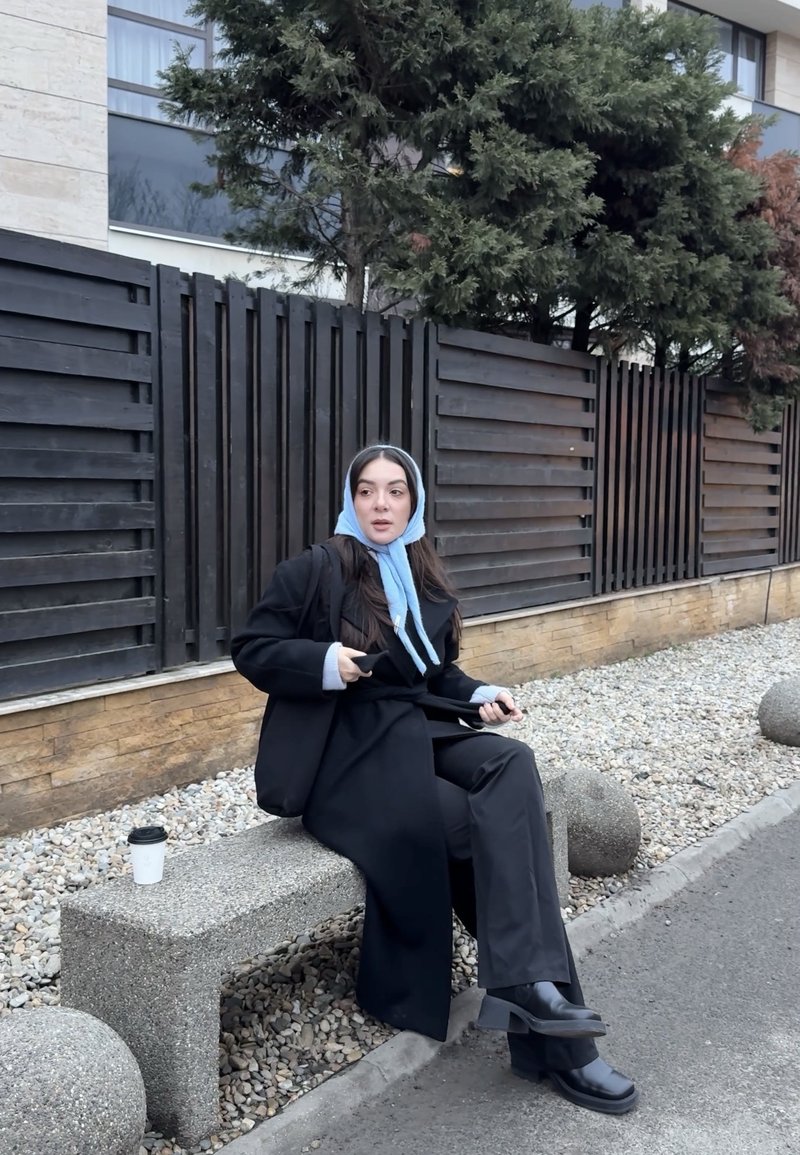 Femme en manteau noir et foulard bleu assise sur un banc en pierre près d'une clôture noire, tenant un téléphone, avec une tasse de café posée à côté d'elle.