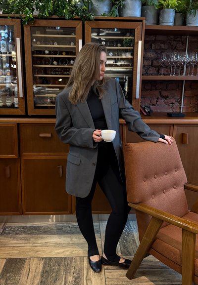 Mujer con blazer gris sosteniendo una taza blanca, parada junto a una silla marrón acolchada en una habitación con armarios de madera y cristalería.