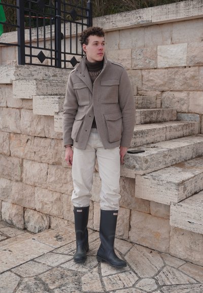 Joven parado junto a unos escalones de piedra, vestido con chaqueta beige, pantalones blancos y botas de lluvia negras, mirando hacia un lado al aire libre.