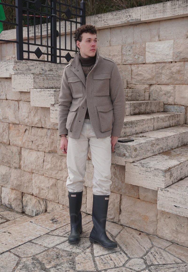 Jeune homme debout près d'escaliers en pierre, portant une veste beige, un pantalon blanc et des bottes de pluie noires, regardant sur le côté en plein air.