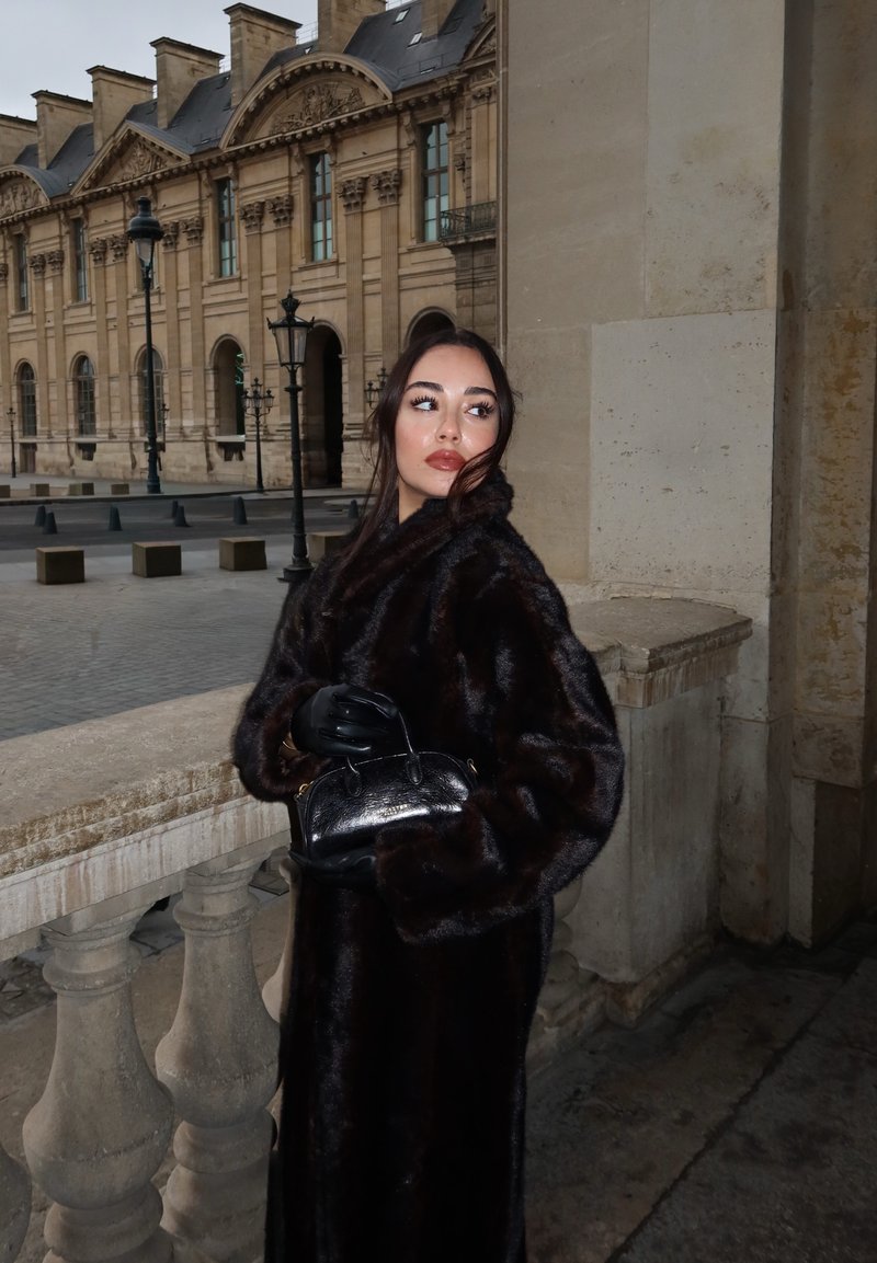 Woman in black fur coat and gloves holding a black handbag, standing by stone railing outside historic building with large windows and street lamps.