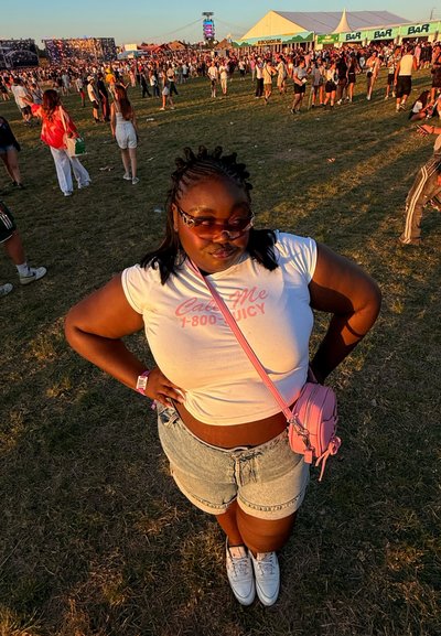Camiseta corta blanca con el texto "Llámae 1-800 JUICY", pantalones cortos de denim claros, bolsa cruzada rosa, gafas de sol, de pie sobre el césped en un festival.