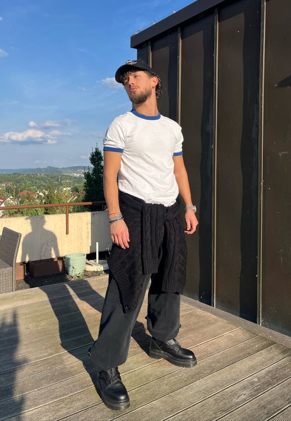 Junger Mann trägt weißes T-Shirt, schwarze Hose und schwarzen Pullover, der in der Taille gebunden ist, und steht auf einem Holzdeck mit Stadtbild im Hintergrund.