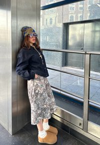 Jeune femme appuyée contre un mur en métal, portant un chapeau camouflage, de grandes lunettes, une veste noire, une jupe à motifs, des chaussettes blanches et des chaussons beiges.