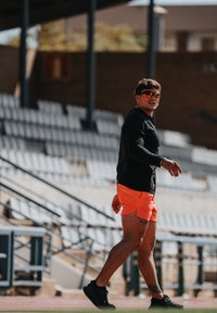 Mann in schwarzem Langarmshirt, leuchtend orangen Shorts und Sonnenbrille, der auf einem Sportfeld mit leeren Stadionplätzen im Hintergrund geht.