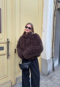 Veste marron duveteuse avec un col montant, associée à un pantalon noir à jambes larges et un petit sac à main noir. En arrière-plan, une porte jaune.