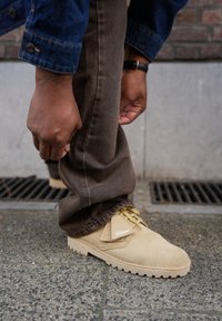 Beige suède schoenen met een gestructureerd oppervlak en een crèmekleurige rubberen zool. Gekoombineerd met donkerbruine broek, die de veters en het logo van de schoen laat zien.