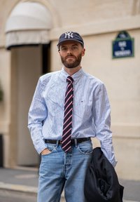 Világoskék és fehér csíkos ing, piros és sötétkék csíkos nyakkendő, világoskék farmernadrág, fekete dzseki a hónalj alatt, sötétkék baseball sapka.