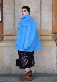 Lühikeste juustega isik helesinisest jakist, musta sädelevat seelikuga, pruunide platvormsandaalide ja tumedate sokkidega, seisab kivimüüri taustal.