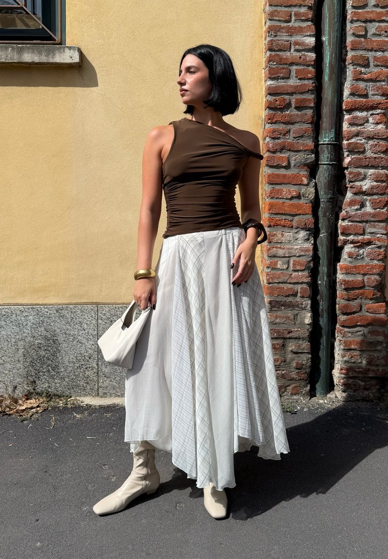 Donna con capelli neri corti, che indossa un top asimmetrico marrone, una lunga gonna bianca a fantasia, stivali beige e tiene in mano una borsa bianca, in piedi accanto a un muro giallo e a un pilastro di mattoni.