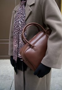 Borsa a mano in pelle marrone con texture liscia e manici arrotondati, tenuta accanto a una sciarpa con motivo leopardato e frange.