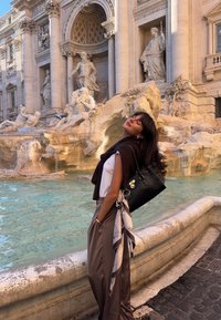 Must käekott hõbedaste detailidega, kombineeritud valge topi ja pruunide pükstega. Kotti ripub hele sall. Taustal on purskkaev.