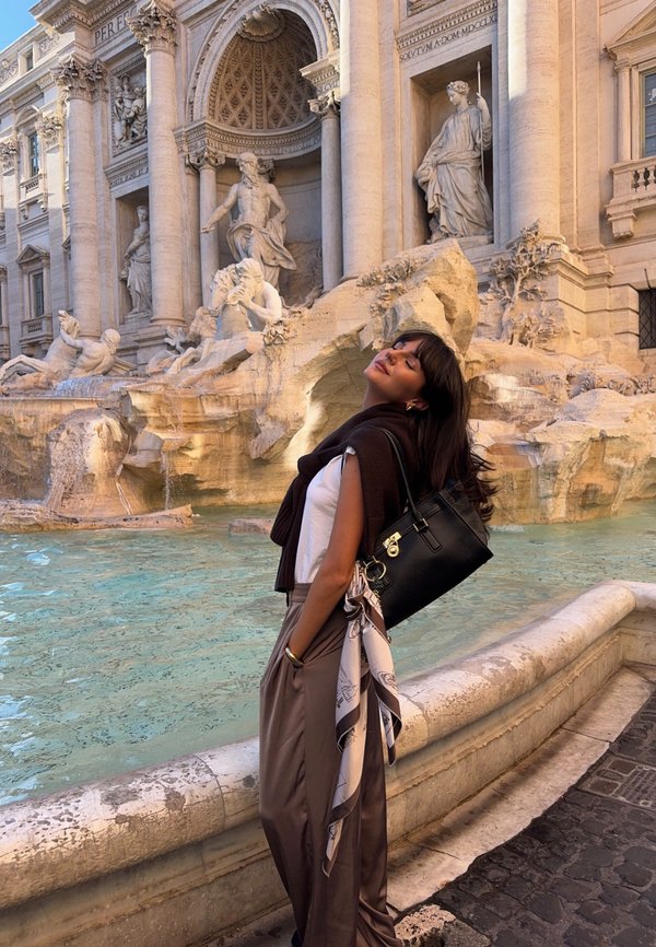 Must käekott hõbedaste detailidega, kombineeritud valge topi ja pruunide pükstega. Kotti ripub hele sall. Taustal on purskkaev.