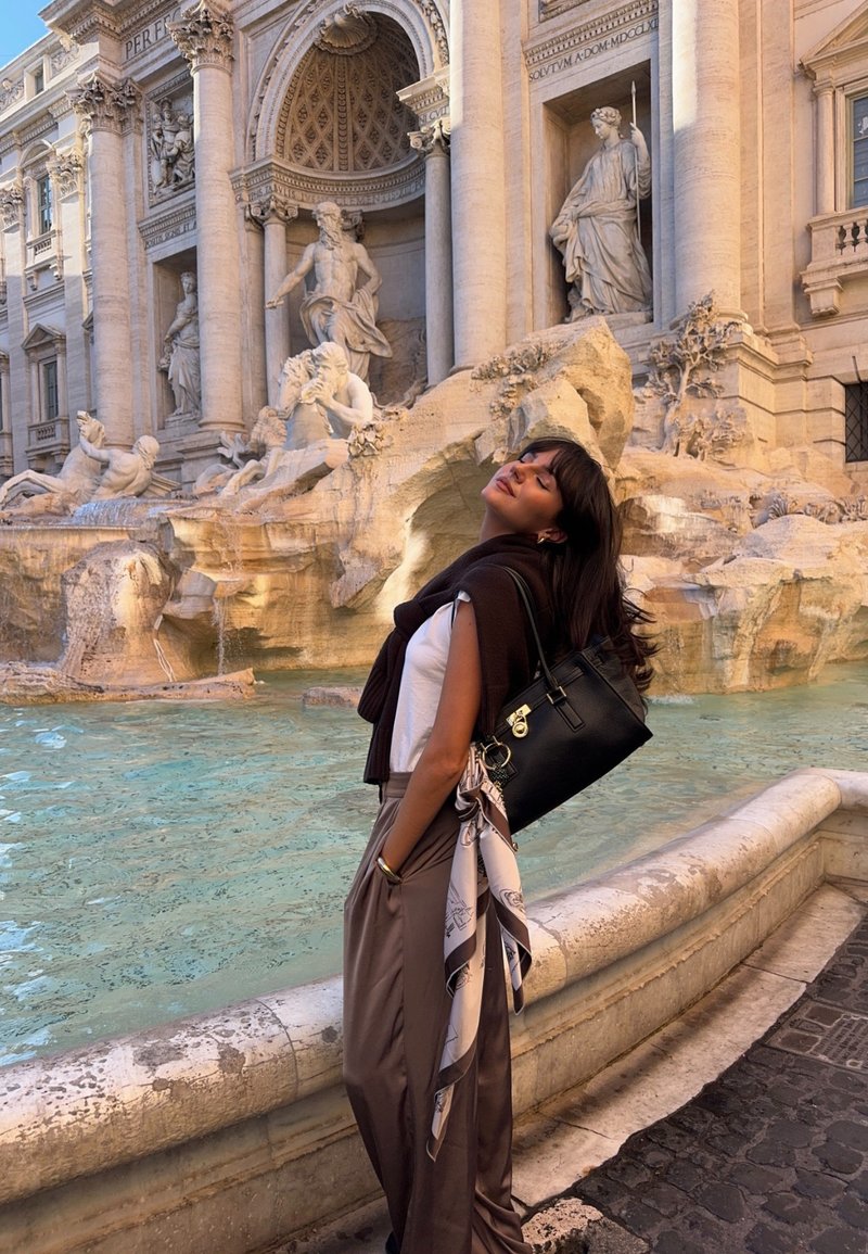 Uma mala preta com ferragens prateadas, combinada com uma blusa branca e calças castanhas. Um lenço claro está pendurado na mala. Fonte de água ao fundo.